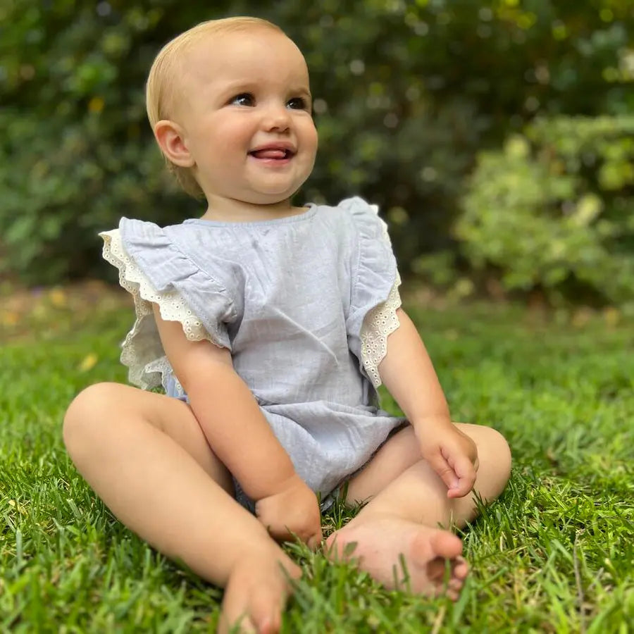 Vignette Megan Bubble Blue baby romper; light blue fabric with lace-trim sleeves, toddler sits barefoot on grass smiling.