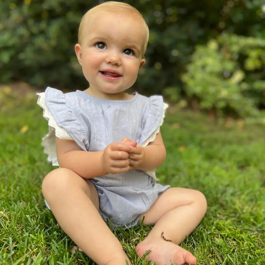 Vignette Megan Bubble Blue baby romper worn by a smiling infant sitting on grass outdoors, blue fabric with lace-trimmed ruffles.