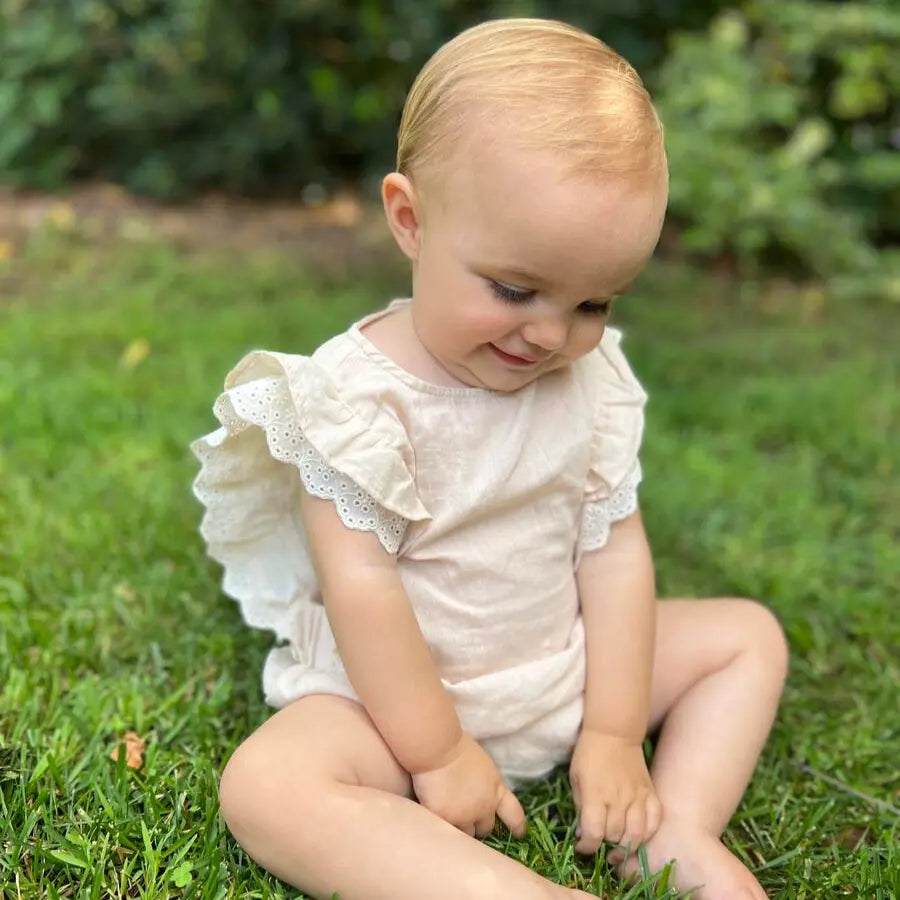Vignette Megan Bubble Cream baby romper in cream fabric with lace flutter sleeves; a baby sits on grass.