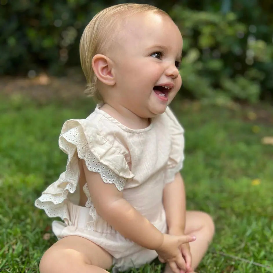 Vignette Megan Bubble Cream baby onesie with lace-trim ruffle sleeves in beige, worn by a smiling toddler on grass.
