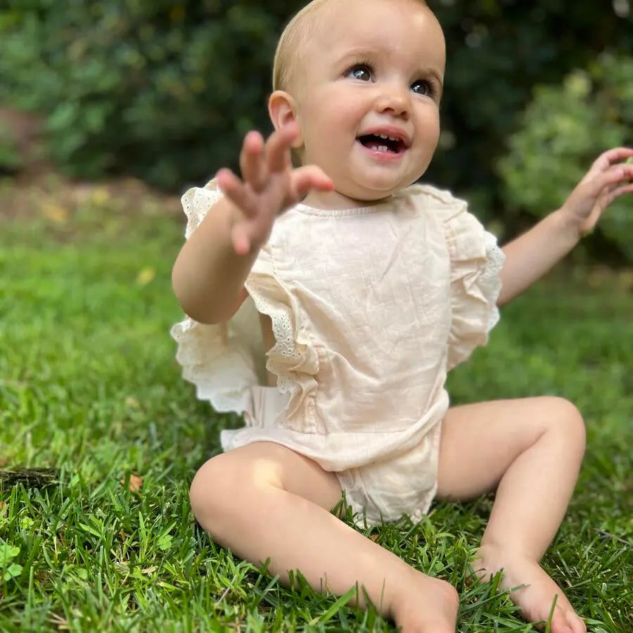 Vignette Megan Bubble Cream baby romper in cream with ruffled sleeves, outdoors on grass as baby reaches.