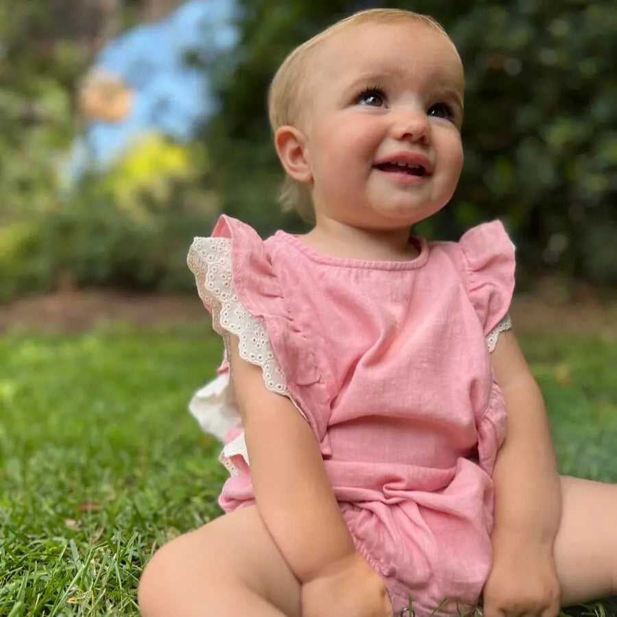 Vignette Megan Bubble Pink baby romper with lace-trim flutter sleeves; smiling baby sits on grass outdoors.