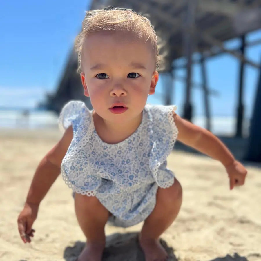Vignette Poppy Bubble romper in blue ditsy floral with lace trim, as a toddler crouches on the sandy beach.