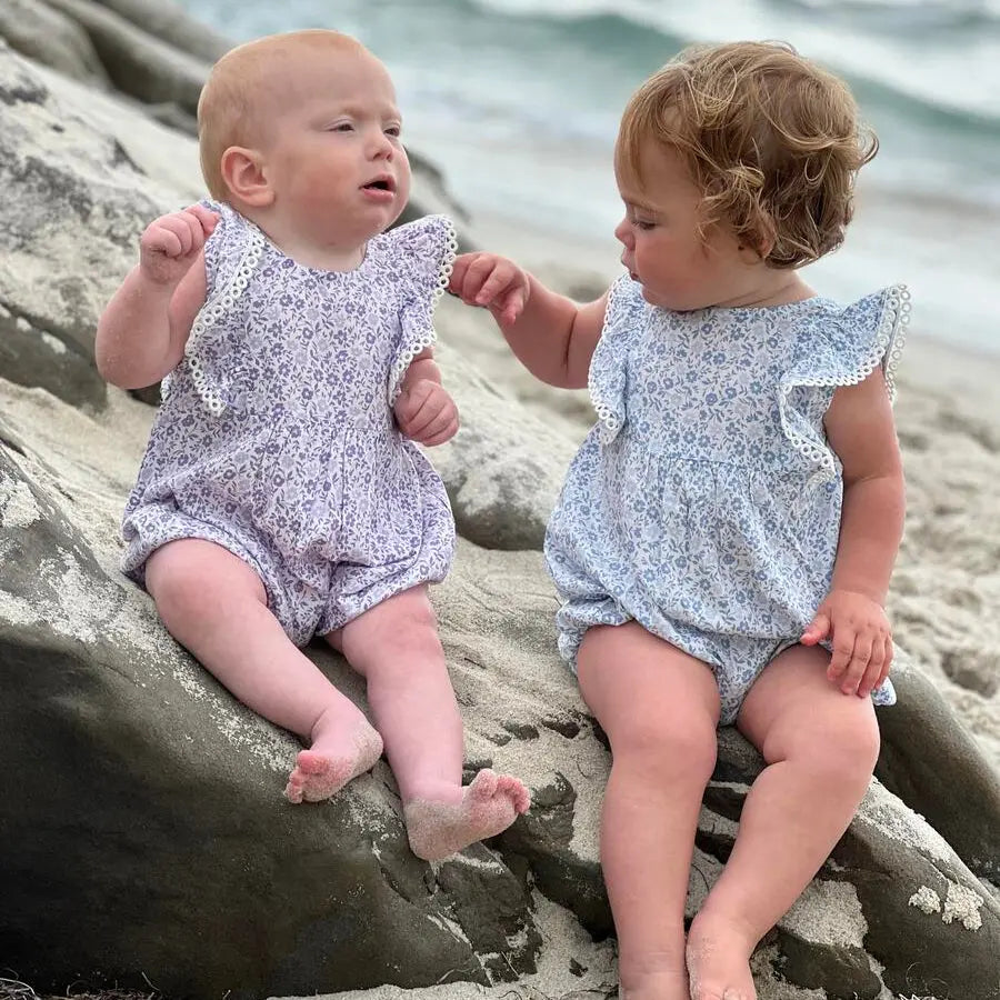 Vignette Poppy Bubble in blue ditsy floral baby romper; two toddlers sit on sea rocks, touching hands.