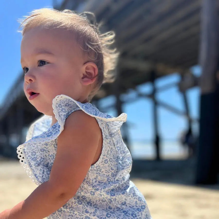 Vignette Poppy Bubble In Blue Ditsy Floral baby romper; blue floral fabric with lace-trim flutter sleeves worn by a toddler at the beach.