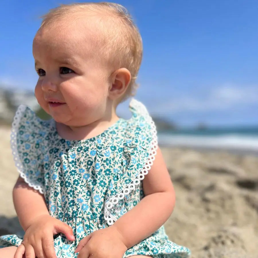 Baby in Vignette Poppy Bubble Teal Ditsy Floral dress with white lace trim sits on a sunlit beach.