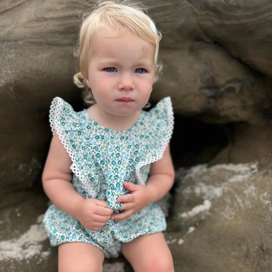Vignette Poppy Bubble teal ditsy floral baby romper with lace trim, worn by toddler sitting on rocks.