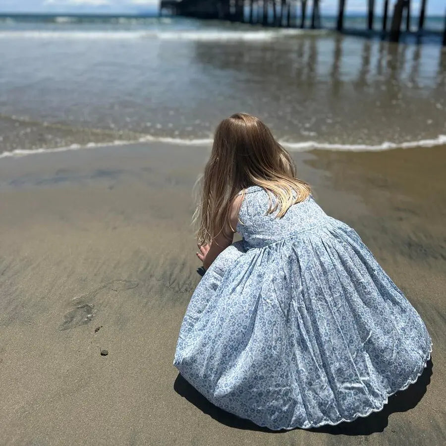 Vignette Stella Dress In Blue Ditsy Floral, child kneels on the beach to reveal the dress's full gathered skirt.