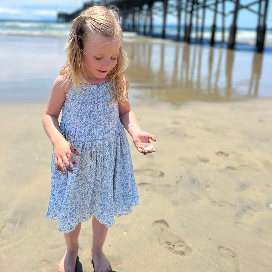 Vignette Stella Dress In Blue Ditsy Floral; toddler girl on the beach, barefoot, holding sand by a pier.