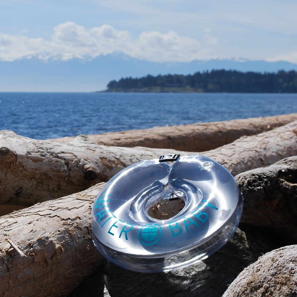 Water Baby Floatie resting on driftwood beside calm sea, silver with blue WATER BABY branding.