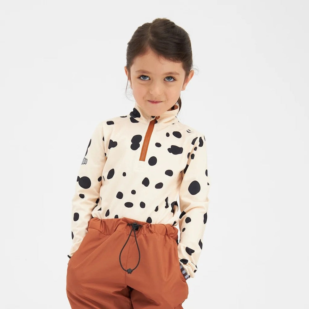 Weedo Puppydo Thermoshirt, cream with black spots and brown zip, paired with rust pants as the girl stands with hands in pockets.