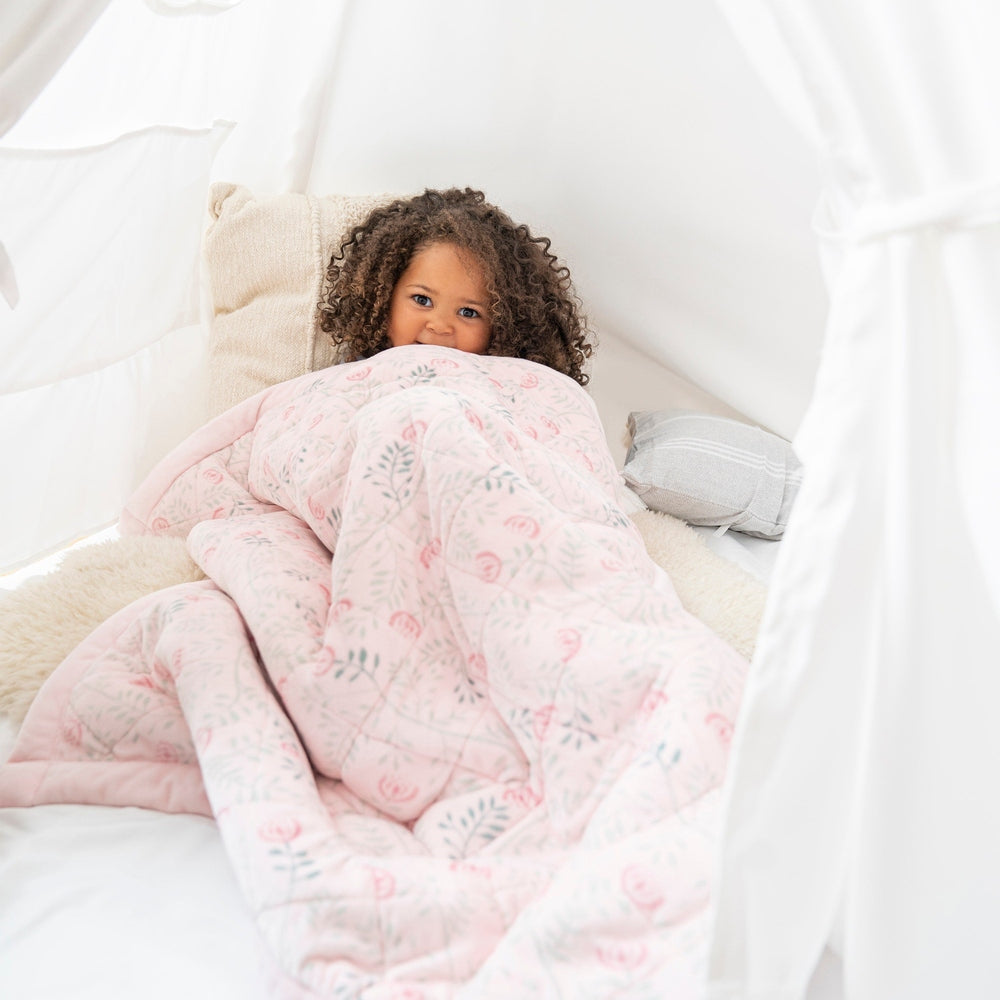 Aden+Anais Embrace Morris Vine weighted toddler blanket, pink floral pattern, wrapped around a smiling child in a white canopy bed