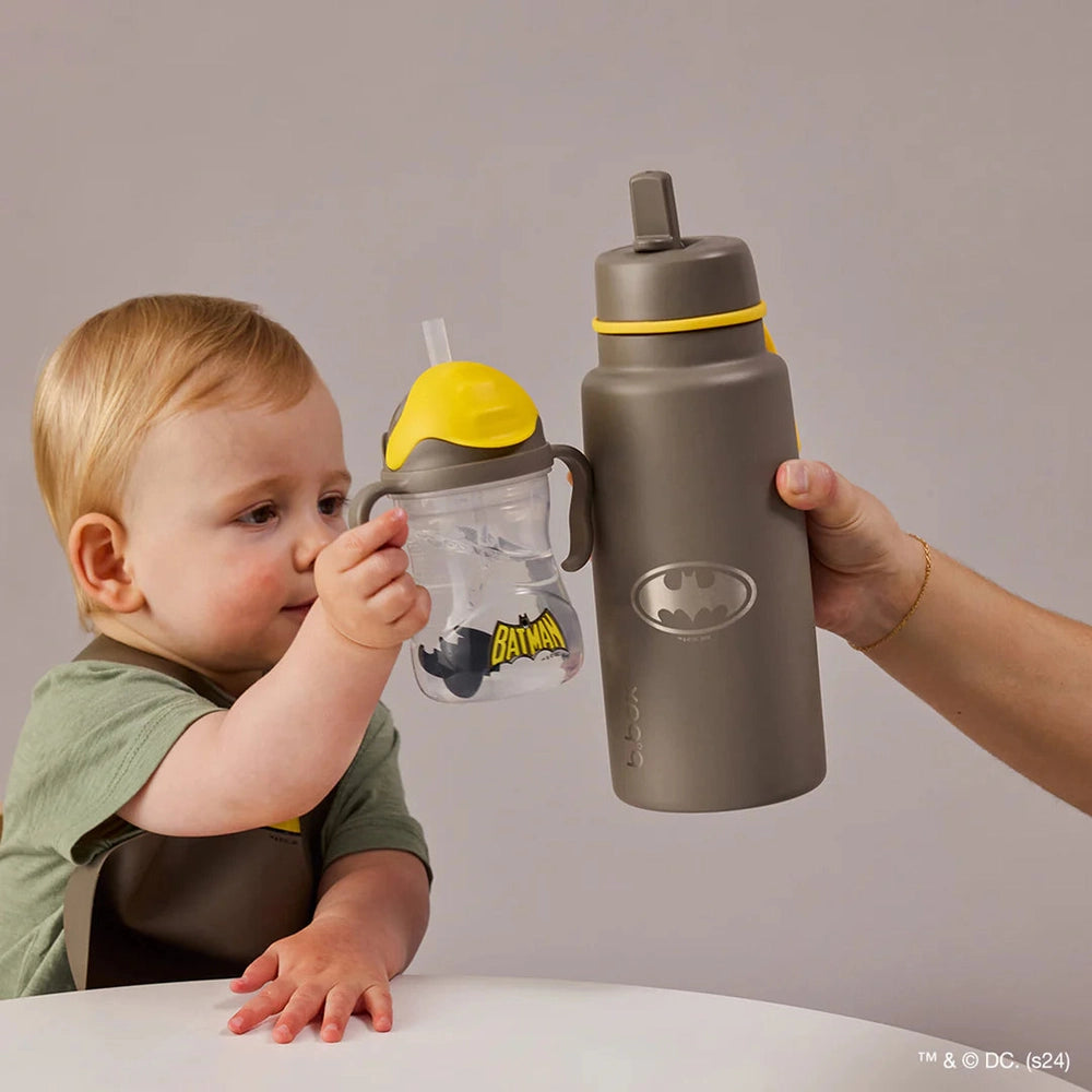 B.box Insulated Flip Top 1L gray water bottle with yellow lid, held by adult beside toddler with Batman cup.