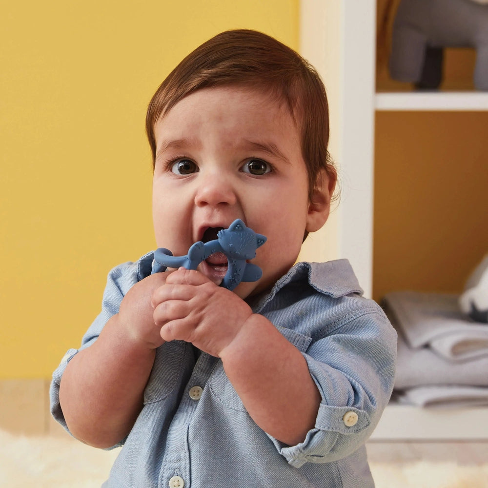 B.Box Wrist Teether worn on baby's wrist; blue silicone dinosaur teether being chewed by a smiling toddler.