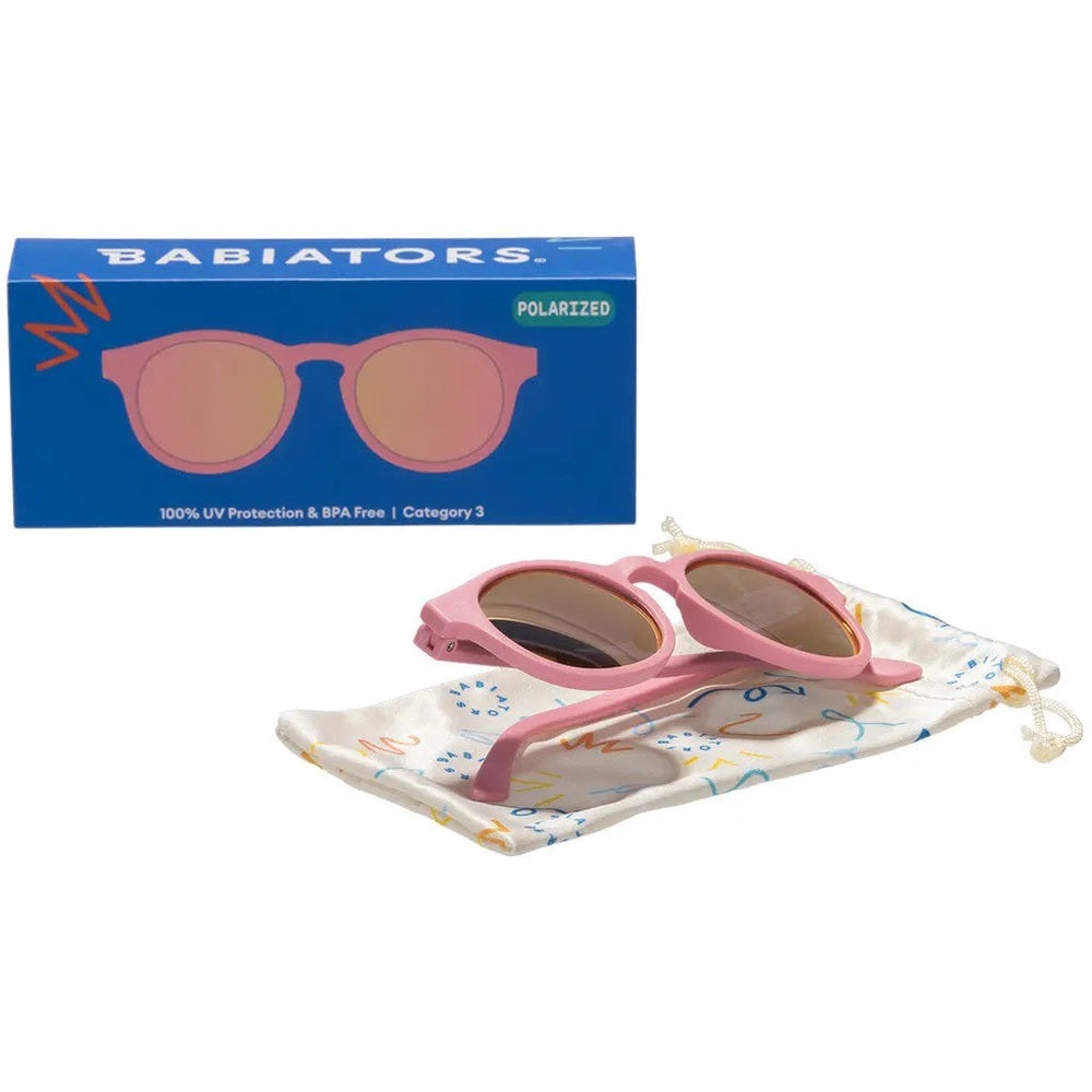 Babiators Polarized Mirrored Sunglasses Keyhole Pretty In Pink, pink frame with mirrored lenses resting on a colorful drawstring pouch.