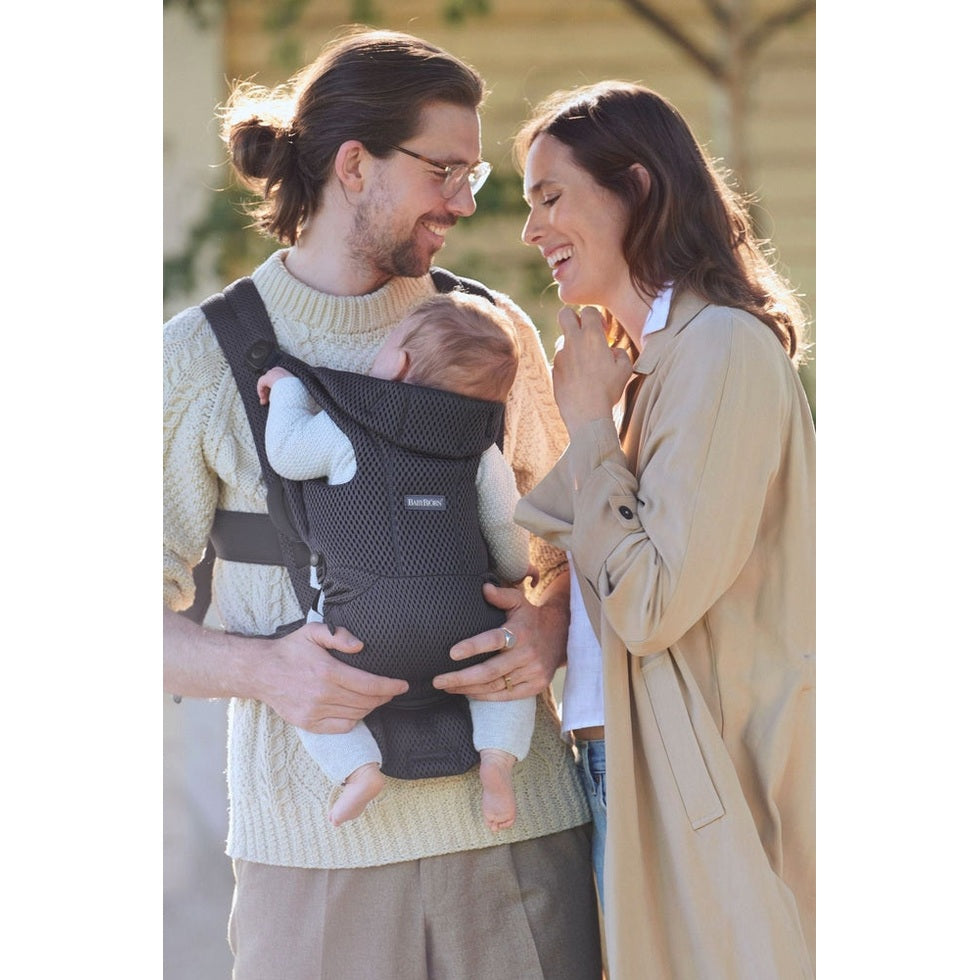 BabyBjorn Baby Carrier Free in dark-grey mesh worn by dad holding baby chest-to-chest while mom smiles outdoors.