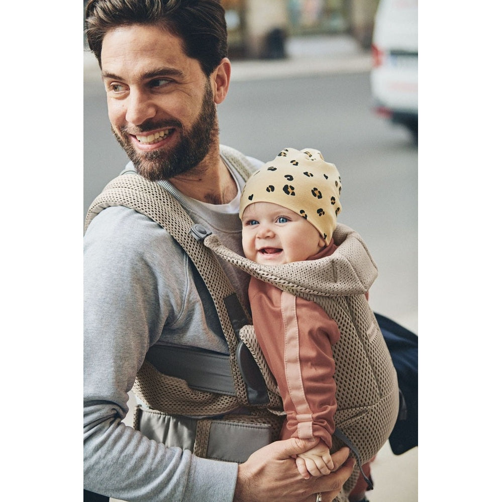 Baby Bjorn Baby Carrier Free in beige worn by dad on front, baby smiling in leopard-print hat.