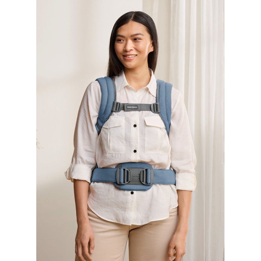 BabyBjorn Baby Carrier Harmony in blue on a smiling woman, showing padded shoulders and wide waist belt.