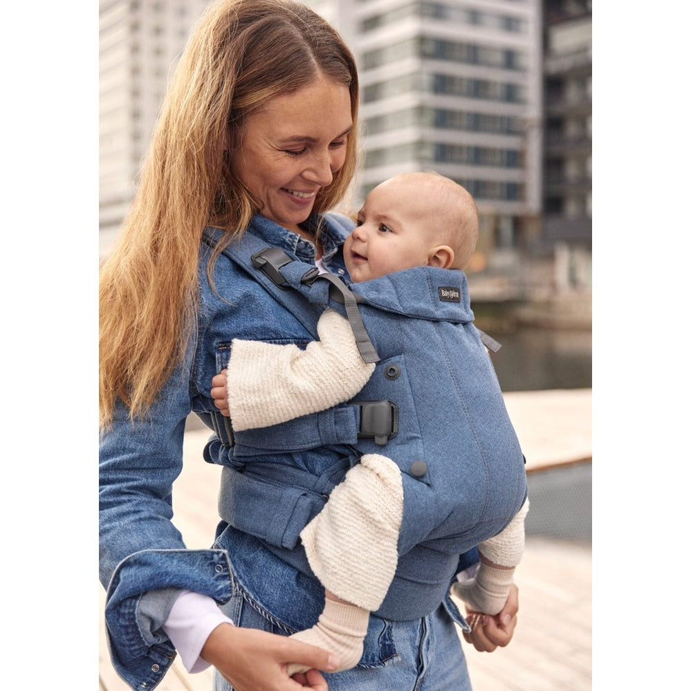 BabyBjorn Baby Carrier Harmony in blue denim worn by smiling mom carrying baby against a city backdrop.