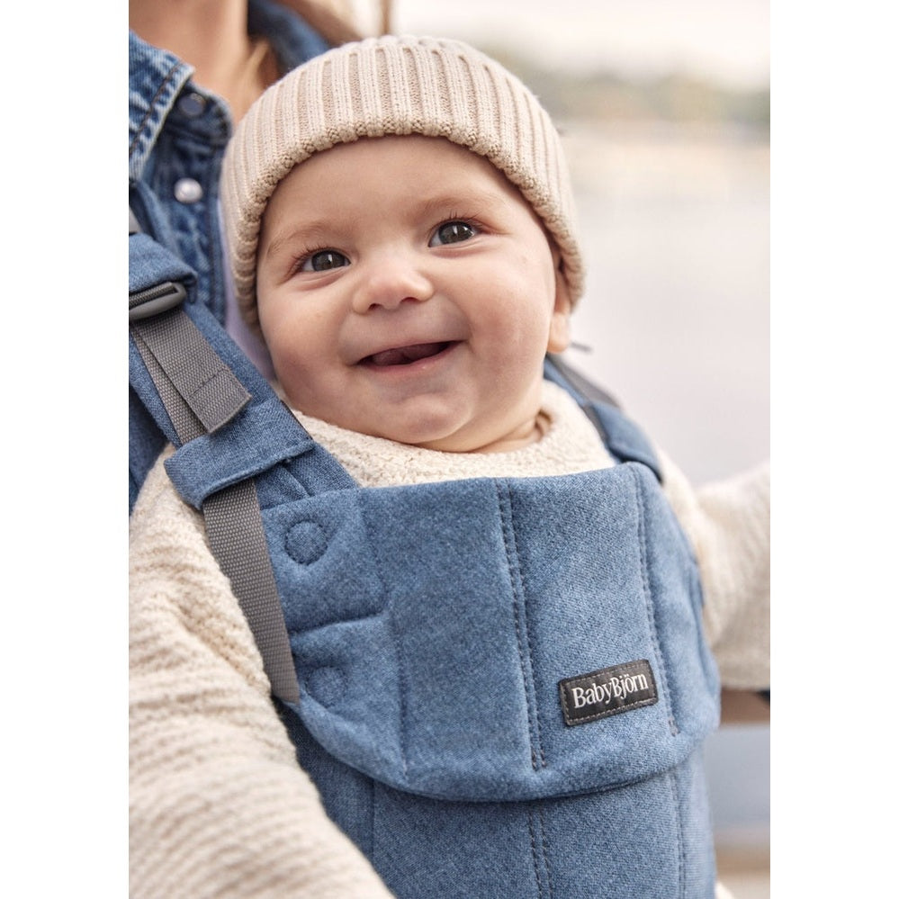 BabyBjorn Harmony baby carrier in blue denim, baby smiling while being securely carried close by a caregiver.