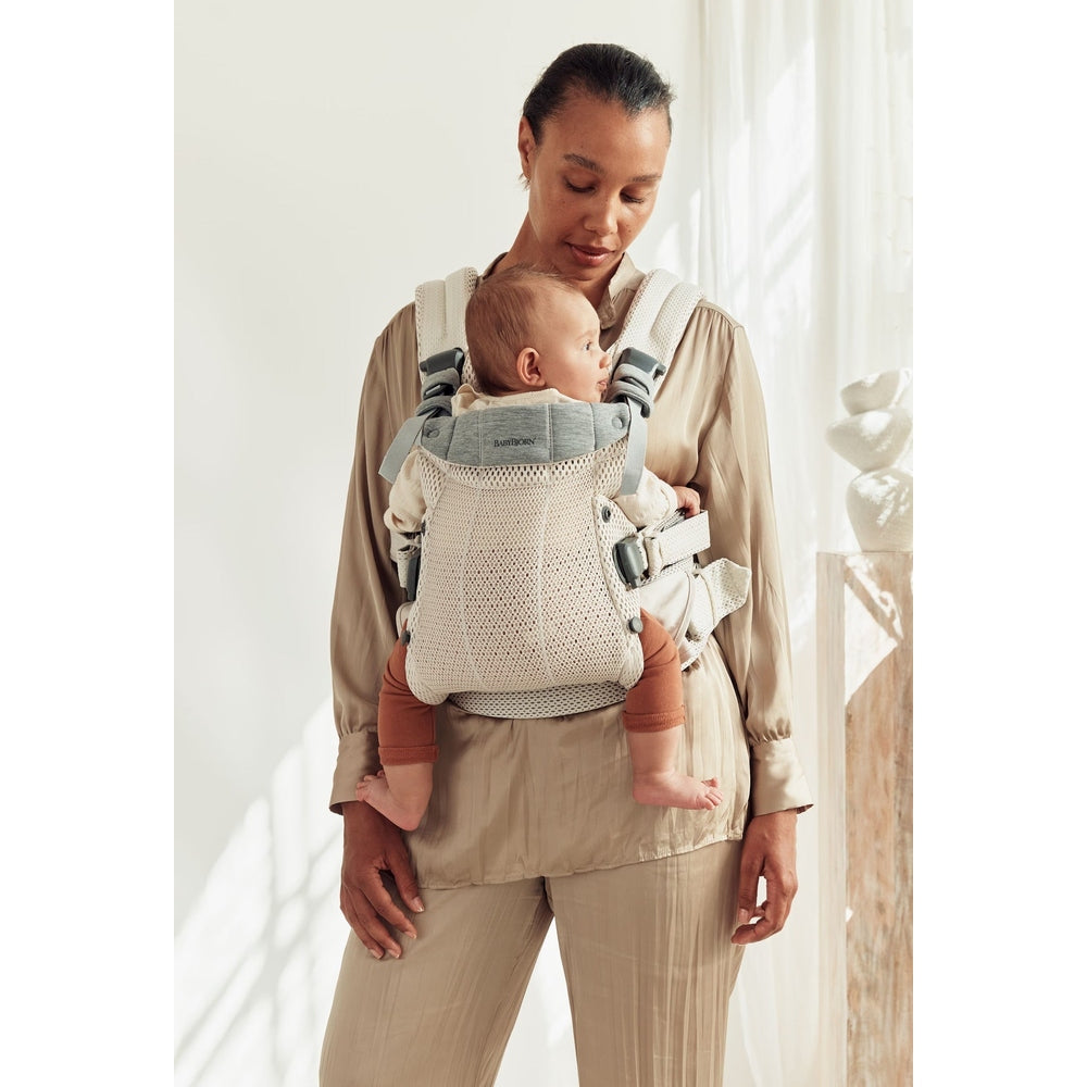 BabyBjorn Harmony Baby Carrier worn by a mother in beige, showing breathable mesh fabric and buckled buckles securing a baby.