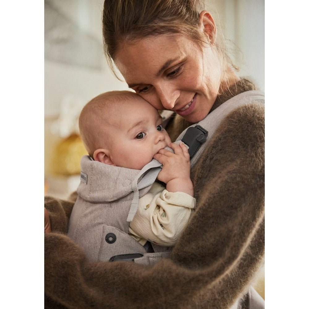BabyBjorn Harmony baby carrier in gray worn by mother, cradling a smiling infant close against her chest.