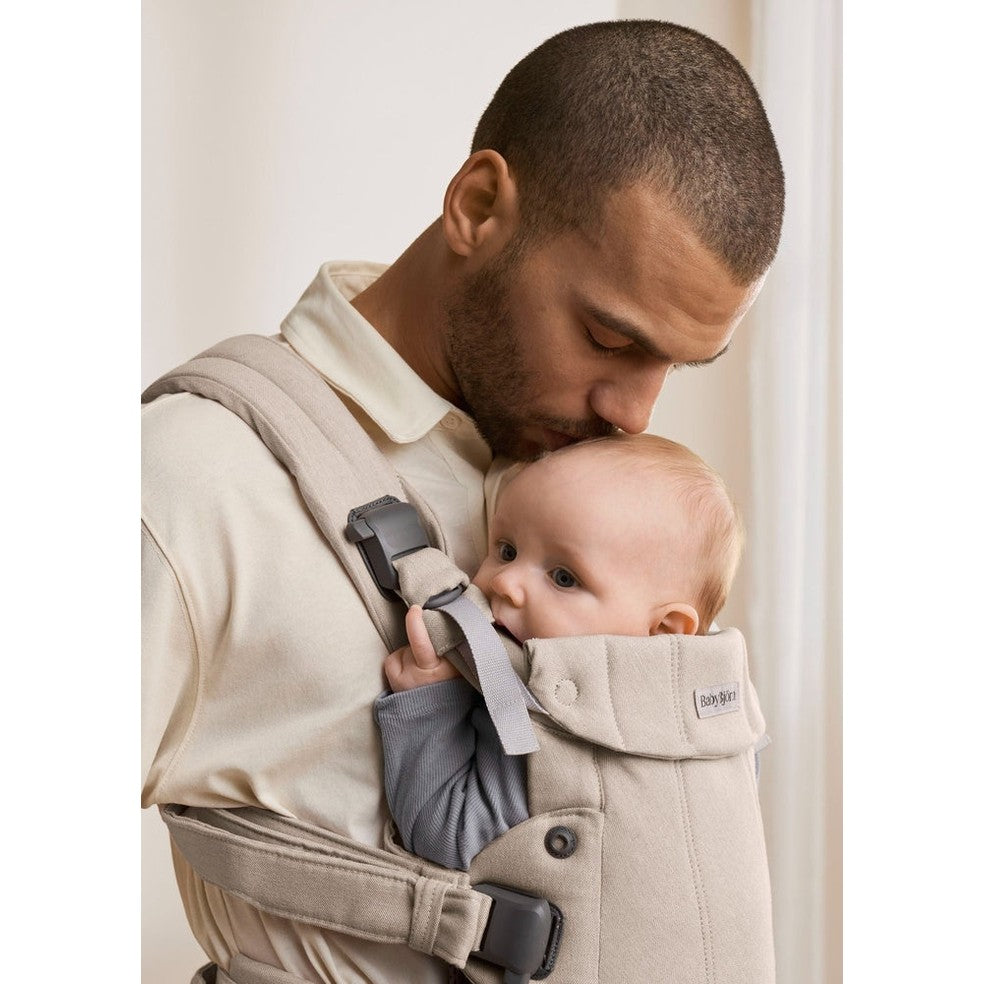 BabyBjorn Baby Carrier Harmony in beige worn by a father holding a baby close with supportive straps and buckles.