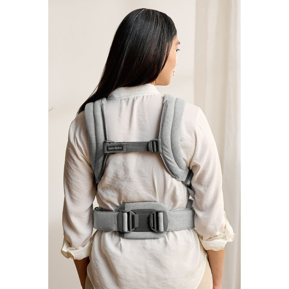 BabyBjorn Baby Carrier Harmony worn by a woman in a light shirt, featuring padded straps and a wide gray waist belt.