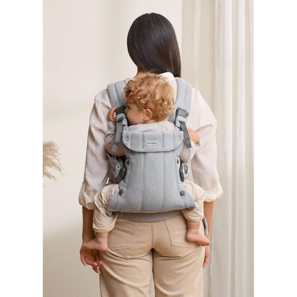 Mother wearing BabyBjorn Baby Carrier Harmony in grey, with padded straps and waist belt, carries toddler on her back.