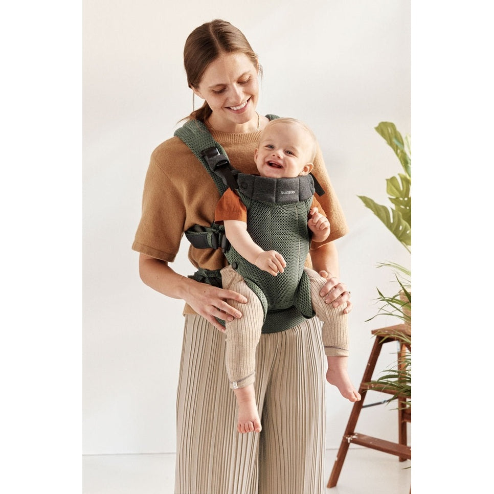 BabyBjorn Harmony baby carrier in green mesh worn by a smiling mom, showing secure waist belt and padded shoulder straps.