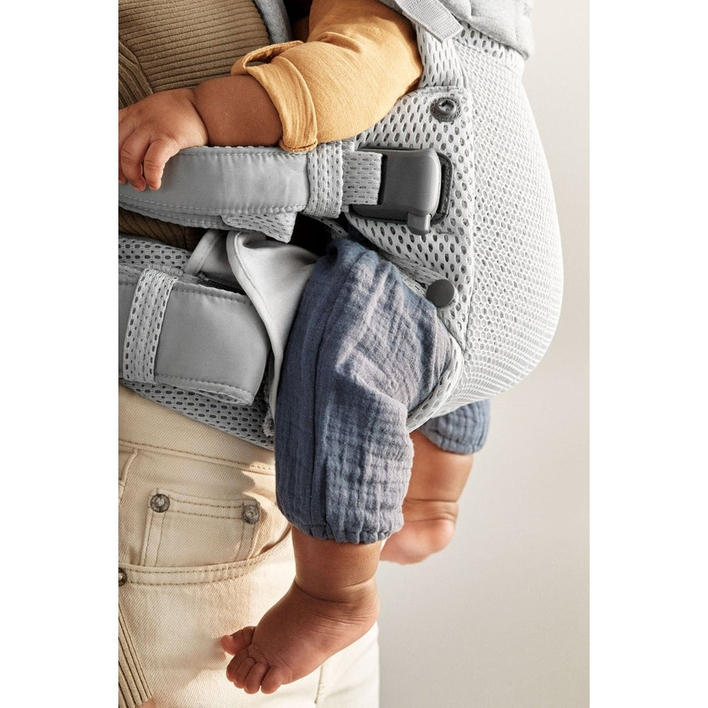BabyBjorn Harmony baby carrier, gray mesh with padded straps, worn by caregiver as baby's legs dangle securely.