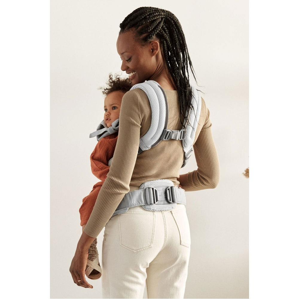 BabyBjorn Harmony baby carrier in light gray worn by smiling mother carrying baby on back with secure straps.
