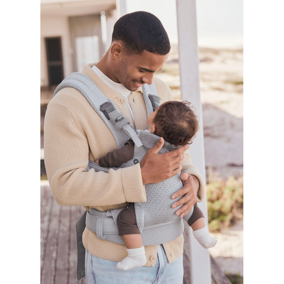 BabyBjorn Harmony baby carrier in light gray with padded straps, as a father gently holds an infant against his chest.