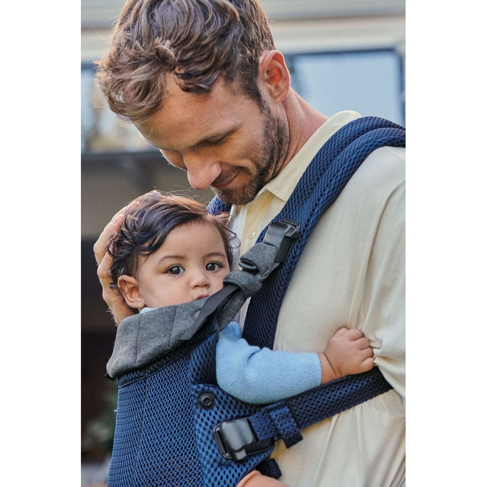 BabyBjorn Harmony baby carrier worn by a father in a beige shirt, baby facing inward and secured with a chest clip.