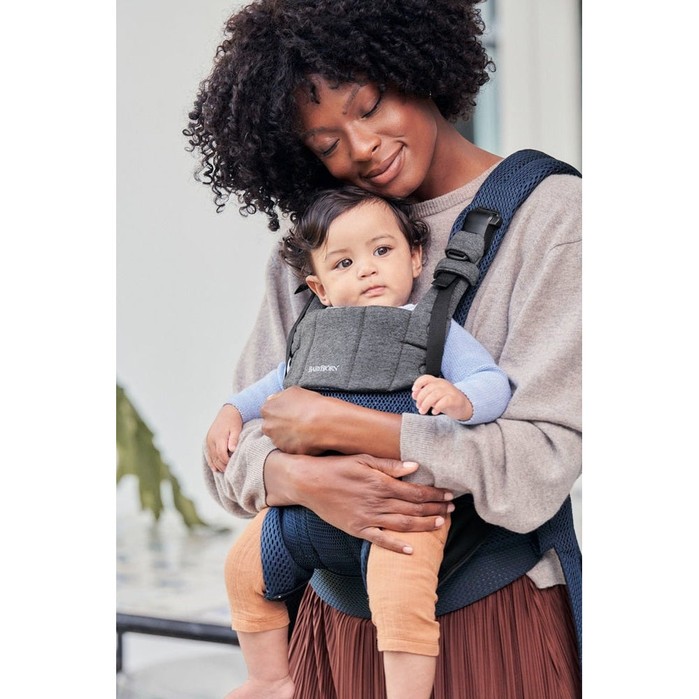 Mother wearing BabyBjorn Baby Carrier Harmony in navy with gray panel, buckled straps, cradling baby securely.