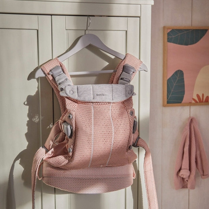 BabyBjorn Baby Carrier Harmony in pink mesh hangs on a hanger, showing padded straps and breathable fabric.