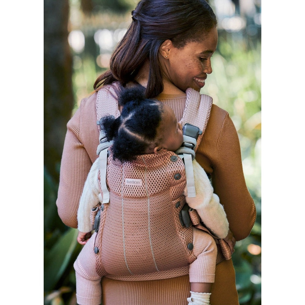 Mother outdoors wearing BABY BJORN Baby Carrier Harmony in blush pink, securely carrying infant on her back.