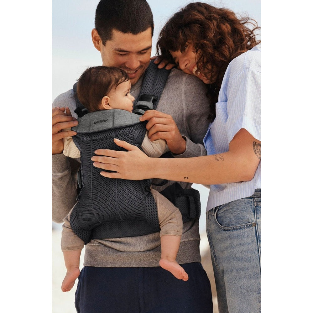 BabyBjorn Harmony baby carrier on beach; parents hold a baby snug against the chest with breathable, ergonomic support.