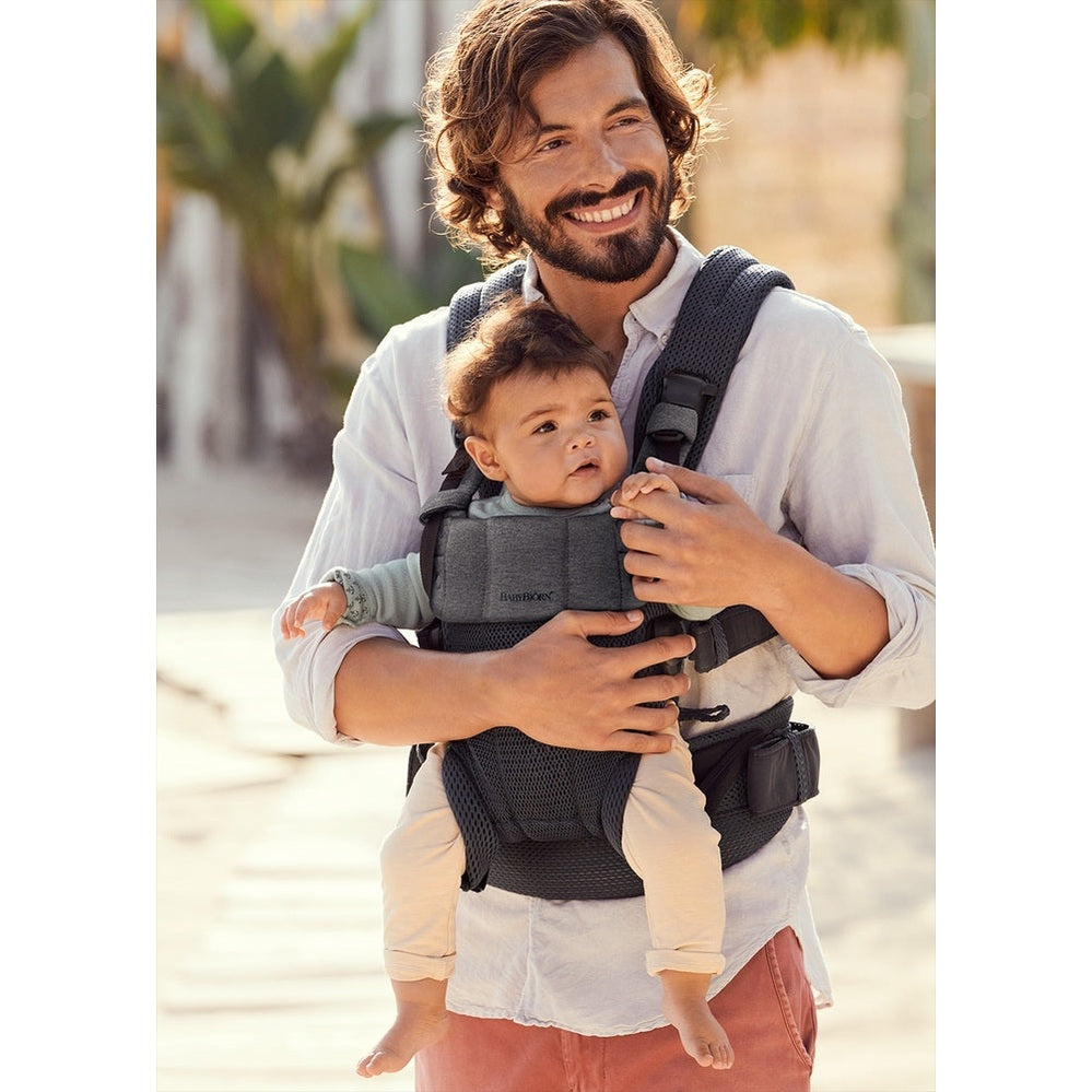 BabyBjorn Baby Carrier Harmony in dark gray worn by a smiling father carrying a baby securely on a sunny walk.