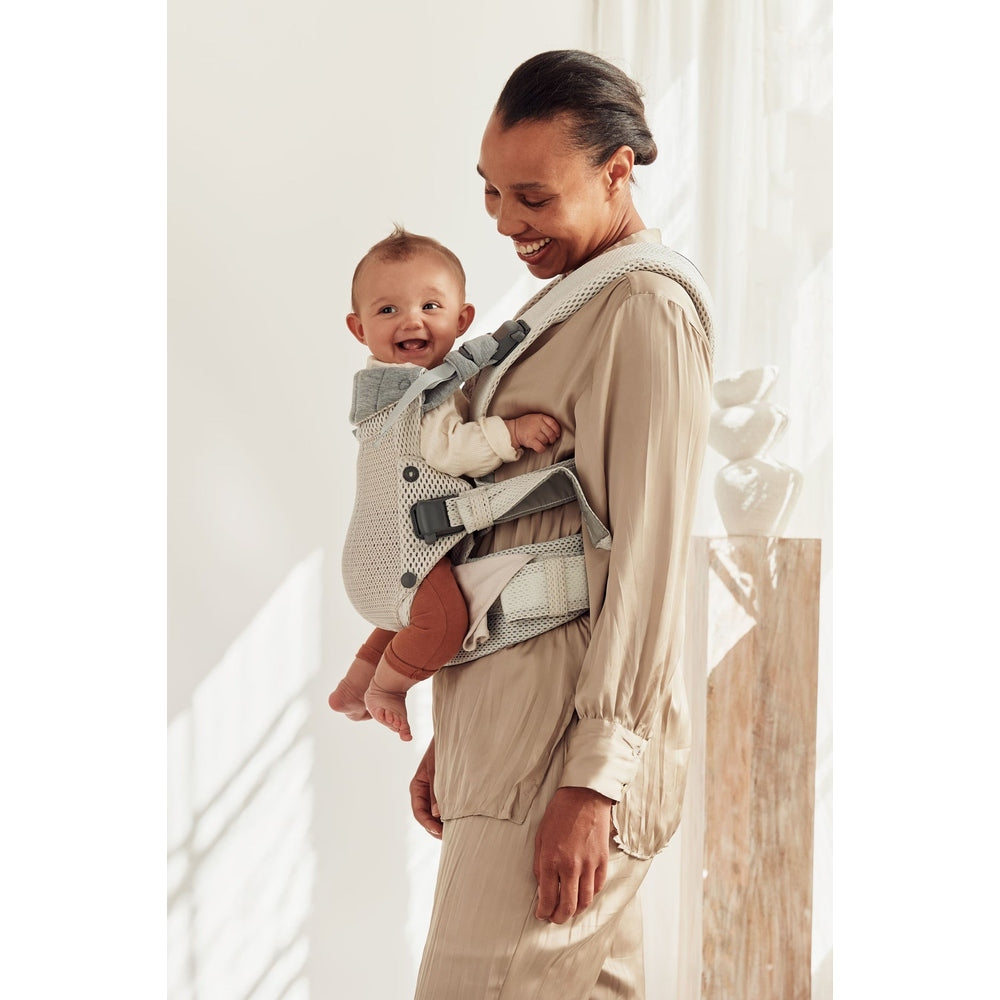 BabyBjorn Harmony baby carrier in beige, parent wearing baby on chest with secure straps, baby smiling.