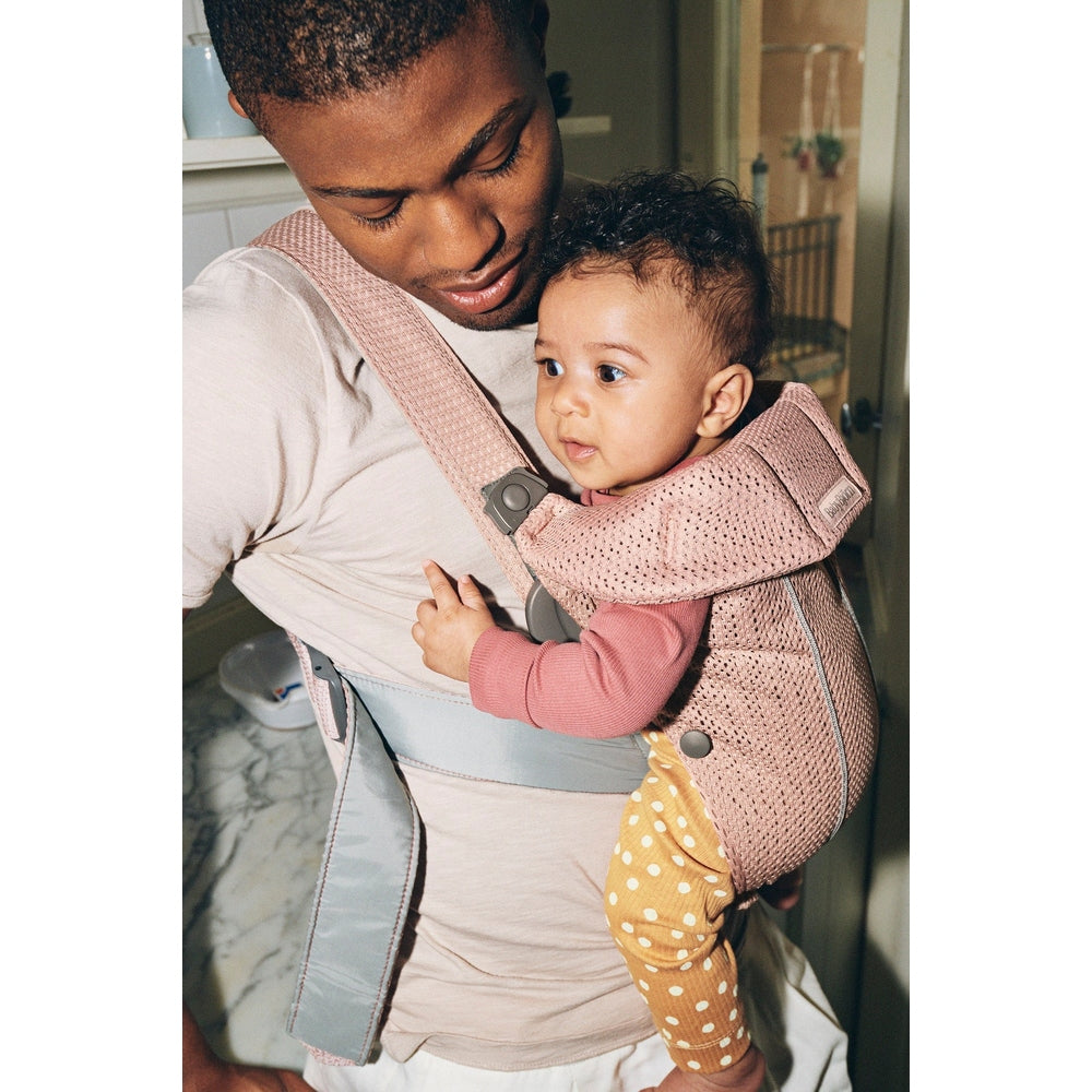 BabyBjorn Baby Carrier Mini in pink mesh, parent buckles chest strap as baby sits facing inward.