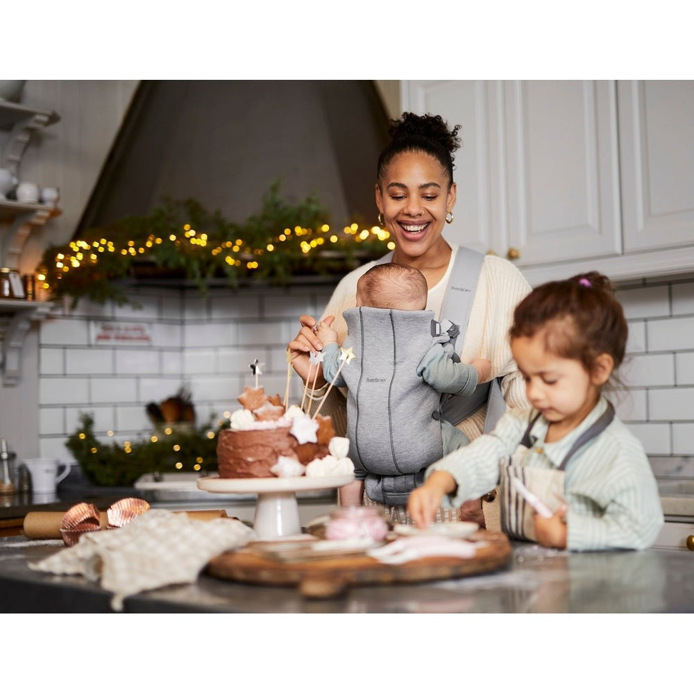 BabyBjorn Baby Carrier Mini in gray worn by smiling mom as she bakes cake with baby secured.