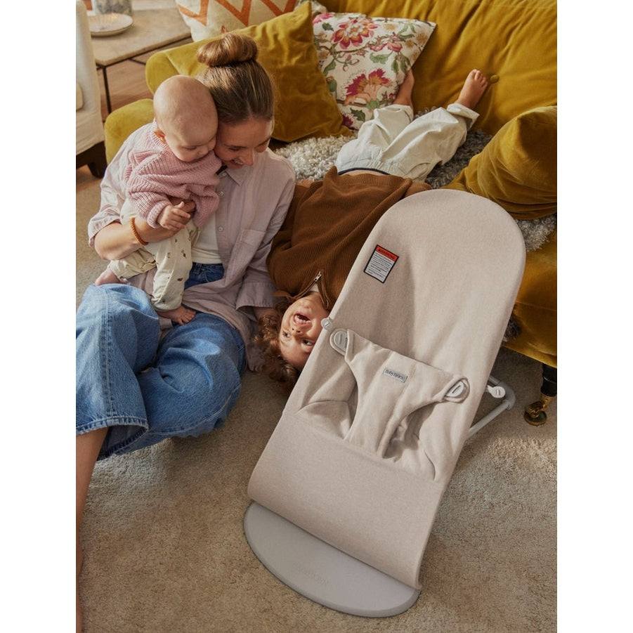 BabyBjorn Bouncer Bliss beige in a cozy living room, mother cuddling a baby while a toddler plays nearby.