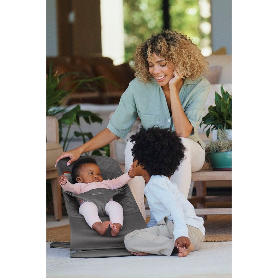 BabyBjorn Bouncer Bliss gray infant bouncer; baby secured in the seat while mom smiles nearby and toddler interacts.