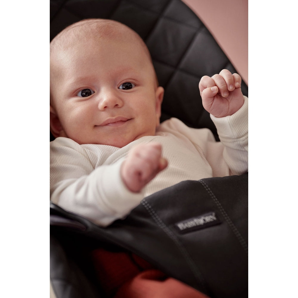 BabyBjorn Bouncer Bliss in black; smiling baby in a white onesie sits securely in the padded seat.