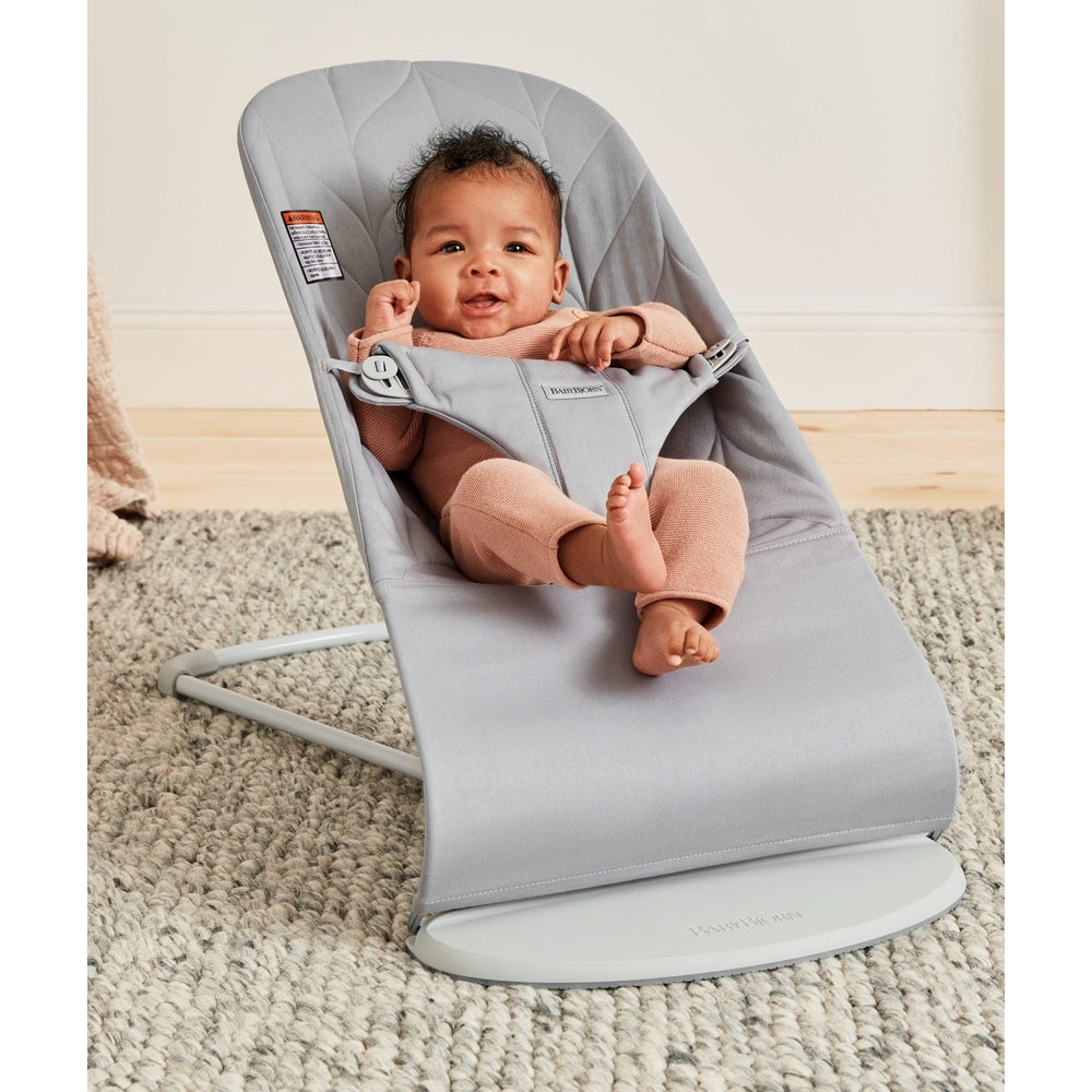 BabyBjorn Bouncer Bliss in soft grey; smiling baby sits securely in the reclined fabric seat using the 5-point harness.