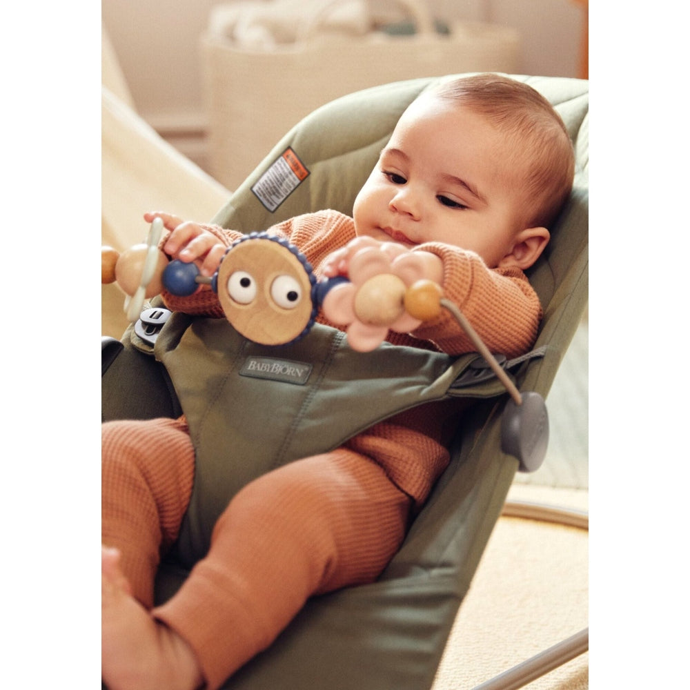 BabyBjorn Bouncer Bliss in green fabric; baby plays with a wooden bead arch while seated and relaxed.