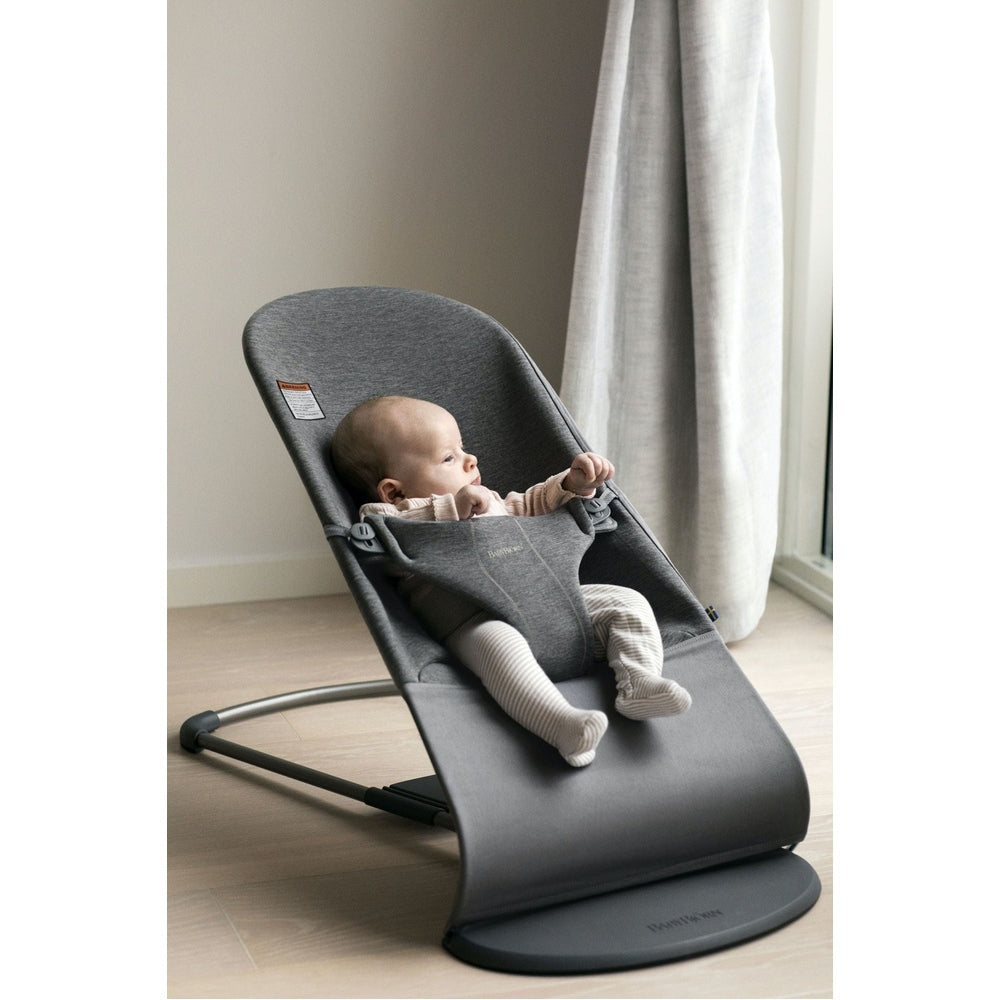 BabyBjorn Bouncer Bliss in gray, a baby relaxing and reaching forward in a bright home setting.