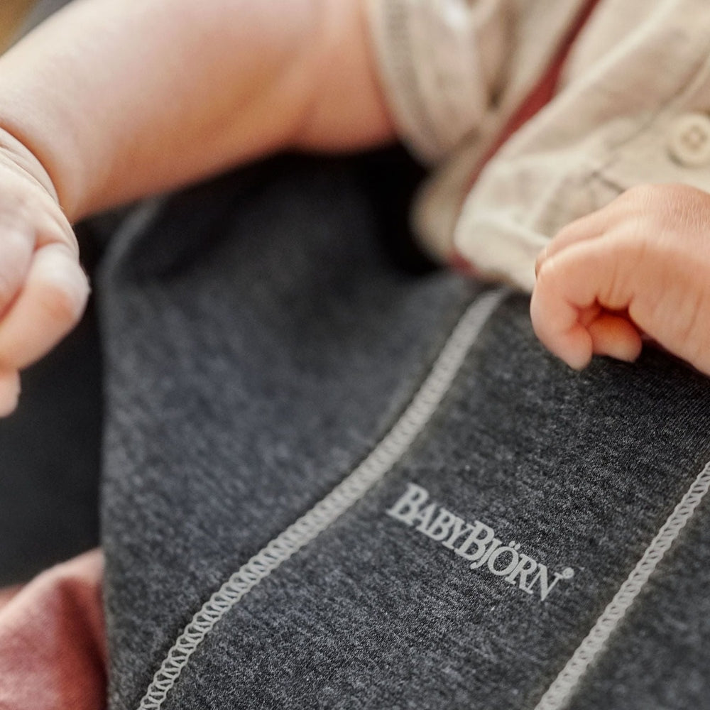 BabyBjorn Bouncer Bliss in dark gray fabric; a baby grips the soft edge, showing plush comfort and sturdy stitching.