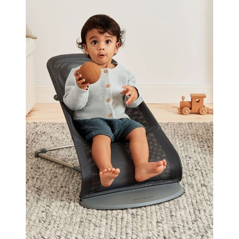 BabyBjorn Bouncer Bliss in black mesh; baby sits and reaches for a textured brown ball.